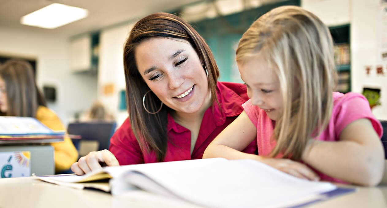 A smiling teacher reading through a workbook alongside a young student at her desk